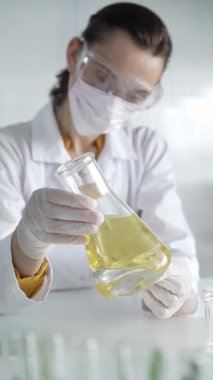 Laboratuvar önlüğü giyen, beyaz eldivenli kadın bilim adamı, laboratuvar ortamında sarı sıvılı Erlenmeyer şişesini tutuyor, portre ve dikey görüntü. Tıp, bilim konsepti.