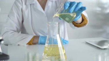 Laboratuvar önlüğü ve mavi koruyucu eldiven giyen bir bilim adamı laboratuarda bir şişeden diğerine sarı bir sıvı boşaltıyor. Tıp ve bilim kavramları.