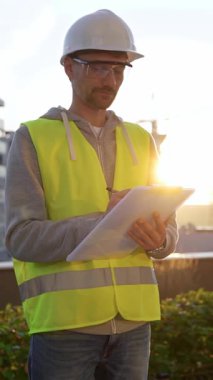 Beyaz kasklı ve güvenlik yeleği olan mühendis, sabahın erken saatlerinde dikey olarak bir inşaat alanını incelerken bir panoya notlar alıyor. Mimarlık ve mühendislik