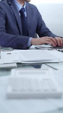 Mavi takım elbiseli iş adamı ofis setindeki cam masada dizüstü bilgisayar, hesap makinesi ve belgelerle çalışıyor. Düşük ışıklandırma, yakın dikey görüntü. İş adamları ve denetim.