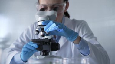 Laboratuvar önlüğü, beyaz koruyucu eldiven ve gözlük giyen kadın bilim adamı modern laboratuvar düzeninde mikroskop kullanıyor, portre görüntüsü. Çaktırmadan ışıklandırma videosu. Bilim ve Tıp.