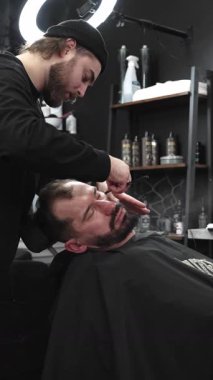 Profesyonel berber müşterilerinin sakallarını ustura ile tıraş ediyor, jöle sürüyor ve modern berber ortamında sıcak havlu kullanıyor, dikey bir bakış açısı var..