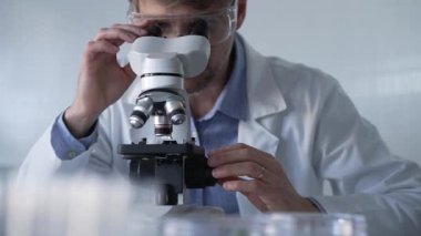 Laboratuvar önlüğü giyen ve koruyucu gözlük takan erkek bilim adamı mikroskop kullanarak modern laboratuvar düzeninde araştırma yapıyor, portre görüntüsü çiziyor. Çaktırmadan ışıklandırma videosu. Bilim ve Tıp Konsepti.
