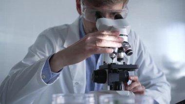 Laboratuvar önlüğü giyen ve koruyucu gözlük takan erkek bilim adamı mikroskop kullanarak modern laboratuvar düzeninde araştırma yapıyor, portre görüntüsü çiziyor. Çaktırmadan ışıklandırma videosu. Bilim ve Tıp Konsepti.