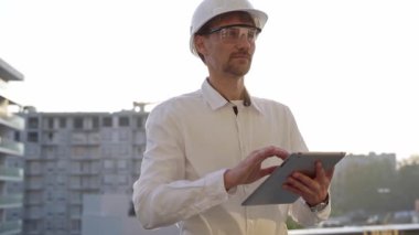 İnşaat mühendisi ya da mimar beyaz gömlekli ve başlıklı adam gün batımında bir inşaat alanını incelerken dijital tablet kullanıyor. Mimarlık ve mühendislik kavramları.