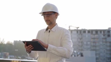İnşaat mühendisi ya da mimar beyaz gömlekli ve başlıklı adam gün batımında bir inşaat alanını incelerken dijital tablet kullanıyor. Mimarlık ve mühendislik kavramları.