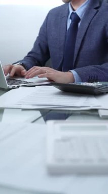 Mavi takım elbiseli iş adamı ofis setindeki cam masada dizüstü bilgisayar, hesap makinesi ve belgelerle çalışıyor. Düşük ışıklandırma, yakın dikey görüntü. İş adamları ve denetim.