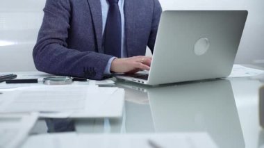 Mavi takım elbiseli iş adamı ofis setindeki cam masada dizüstü bilgisayar, hesap makinesi ve belgelerle çalışıyor. Düşük ışıklandırma, yakın görüş. İş adamları ve denetim kavramı.