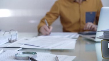 Hesap makinesi, büyüteç, kalem ve belgeler muhasebecilerin masasında. Sarı tişörtlü erkek muhasebeci, arka planda belgelerle çalışıyor. İş ve denetim kavramı.