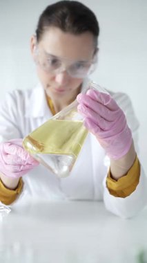 Laboratuvar önlüğü, pembe eldiven ve koruyucu gözlük takan kadın bilim adamı, laboratuvar ortamında, portre ve dikey manzarada sarı sıvı içeren Erlenmeyer matarası tutuyor. Tıp, bilim..