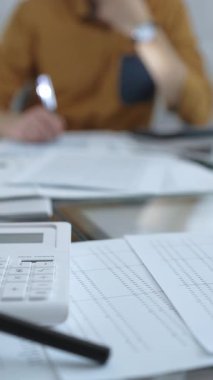Hesap makinesi, büyüteç, kalem ve belgeler muhasebecilerin masasında. Sarı tişörtlü erkek muhasebeci, arka planda belgelerle çalışıyor. Dikey görünüm. İş ve denetim.