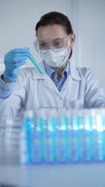 Palto, mavi tıbbi eldiven ve koruyucu gözlük giyen kadın laboratuvar çalışanı veya bilim adamı modern laboratuarda mavi sıvıyla test tüpünü inceliyor. Çaktırmadan ışıklandırma videosu. Bilim ve Tıp.