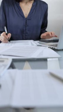 Mavi elbise giyen iş kadını profesyonel ofis ortamında dizüstü bilgisayar, hesap makinesi ve belgelerle çalışıyor. Düşük ışıklandırma, yakın dikey görüntü. İş adamları, denetim kavramı.
