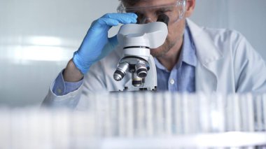 Mavi eldiven giyen, koruyucu gözlüklü erkek araştırmacı modern laboratuarda mikroskop kullanıyor, portre görüntüsü. Gösterişsiz ışıklandırma fotoğrafı. Bilim ve Tıp kavramları.