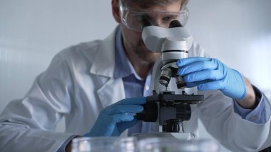 Mavi eldiven giyen, koruyucu gözlüklü erkek araştırmacı modern laboratuarda mikroskop kullanıyor, portre görüntüsü. Gösterişsiz ışıklandırma fotoğrafı. Bilim ve Tıp kavramları.