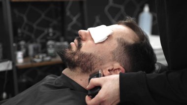 Profesyonel berber, müşterinin sakalını tıraş etmek için ustalıkla elektrikli jilet kullanır. Zarif ve modern bir berber dükkanında titizlikle bakımı sağlar..