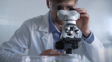 Laboratuvar önlüğü giyen ve koruyucu gözlük takan erkek bilim adamı mikroskop kullanarak modern laboratuvar düzeninde araştırma yapıyor, portre görüntüsü çiziyor. Gösterişsiz ışıklandırma fotoğrafı. Bilim ve Tıp Konsepti.
