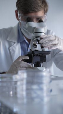 Laboratuvar önlüğü, beyaz eldiven, tıbbi maske ve koruyucu gözlük takan bilim adamı mikroskop kullanarak modern laboratuarda araştırma yapıyor, portre görüntüsü alıyor. Gösterişsiz ışıklandırma fotoğrafı. Bilim ve Tıp.