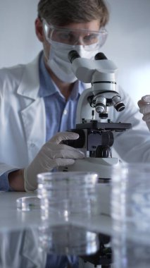 Laboratuvar önlüğü, beyaz eldiven, tıbbi maske ve koruyucu gözlük takan bilim adamı mikroskop kullanarak modern laboratuarda araştırma yapıyor, portre görüntüsü alıyor. Gösterişsiz ışıklandırma fotoğrafı. Bilim ve Tıp.