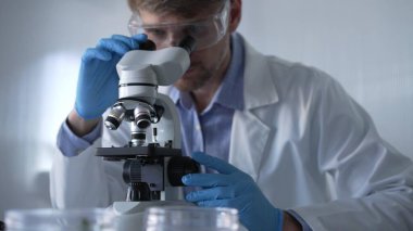 Mavi eldiven giyen, koruyucu gözlüklü erkek araştırmacı modern laboratuarda mikroskop kullanıyor, portre görüntüsü. Gösterişsiz ışıklandırma fotoğrafı. Bilim ve Tıp kavramları.