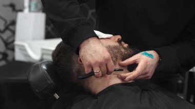 Profesyonel berber müşterisinin sakalını jiletle tıraş eder, tıraş jölesi sürer ve modern bir berber dükkanında tıraş olmak için sıcak havlu kullanır.