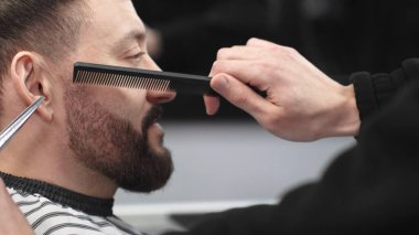 Profesyonel berber, müşterinin sakalını makas ve tarak ile tarayıp keserek modern bir berber dükkanında şık ve titiz bir tımar deneyimi sağlar.