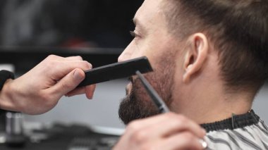 Profesyonel berber, müşterinin sakalını makas ve tarak ile tarayıp keserek modern bir berber dükkanında şık ve titiz bir tımar deneyimi sağlar.