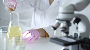 Laboratuvar önlüğü ve pembe koruyucu eldiven giyen bir bilim adamı laboratuarda bir şişeden diğerine sarı yağlı bir sıvı döküyor. Tıp ve Bilim.