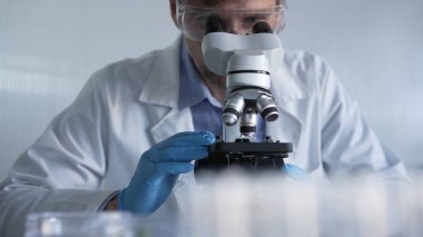 Mavi eldiven giyen, koruyucu gözlüklü erkek araştırmacı modern laboratuarda mikroskop kullanıyor, portre görüntüsü. Gösterişsiz ışıklandırma fotoğrafı. Bilim ve Tıp kavramları.