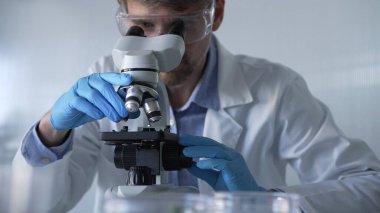 Mavi eldiven giyen, koruyucu gözlüklü erkek araştırmacı modern laboratuarda mikroskop kullanıyor, portre görüntüsü. Gösterişsiz ışıklandırma fotoğrafı. Bilim ve Tıp kavramları.