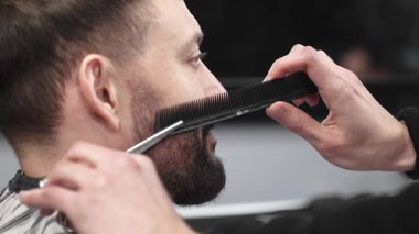 Profesyonel berber, müşterinin sakalını makas ve tarak ile tarayıp keserek modern bir berber dükkanında şık ve titiz bir tımar deneyimi sağlar.