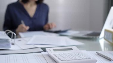 Beyaz hesap makinesi, kalem ve belgeler muhasebecilerin masasında yatıyor. Mavi elbise giyen iş kadını arka planda belgelerle çalışıyor. İş ve denetim kavramı.