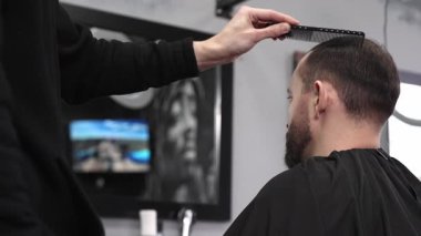 Berber dükkanında şık bir saç kesimine hazırlanırken bir adamın saçını tarayan kuaför aynada görünen bir berber direğinin yansımasıyla canlı bir atmosfer yaratıyor.