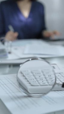 Büyüteç, beyaz hesap makinesi, kalem ve belgeler muhasebecilerin masasında yatıyor. Mavi elbiseli iş kadını arka planda, dikey görünümlü belgelerle çalışıyor. İş ve denetim.
