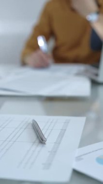 Hesap makinesi, büyüteç, kalem ve belgeler muhasebecilerin masasında. Sarı tişörtlü erkek muhasebeci, arka planda belgelerle çalışıyor. Dikey görünüm. İş ve denetim.