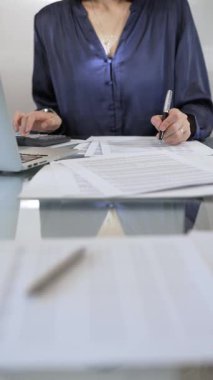 Mavi elbise giyen iş kadını profesyonel ofis ortamında dizüstü bilgisayar, hesap makinesi ve belgelerle çalışıyor. Düşük ışıklandırma, yakın dikey görüntü. İş adamları, denetim kavramı.