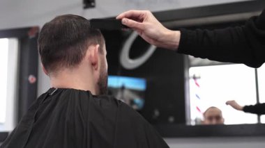 Berber dükkanında şık bir saç kesimine hazırlanırken bir adamın saçını tarayan kuaför aynada görünen bir berber direğinin yansımasıyla canlı bir atmosfer yaratıyor.