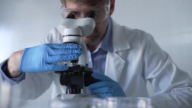 Mavi eldiven giyen, koruyucu gözlüklü erkek araştırmacı modern laboratuarda mikroskop kullanıyor, portre görüntüsü. Çaktırmadan ışıklandırma videosu. Bilim ve Tıp kavramları.