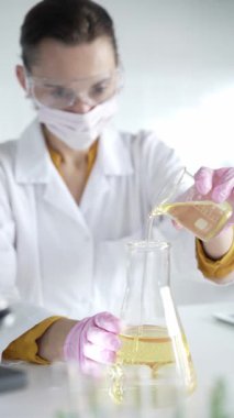 Laboratuvar önlüğü, pembe eldiven ve koruyucu gözlük takan kadın bilim adamı deney şişesinden Erlenmeyer şişesine laboratuvar, portre ve dikey görüntüye sarı bir sıvı döküyor. Tıp, bilim..