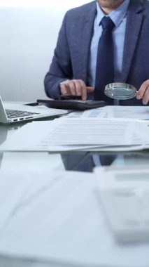Mavi takım elbiseli bir işadamı büyüteçle mali raporları analiz ediyor ve profesyonel ofis ortamında hesap makinesi kullanıyor. Düşük ışıklandırma, yakın dikey görüntü. İş adamları, denetim.