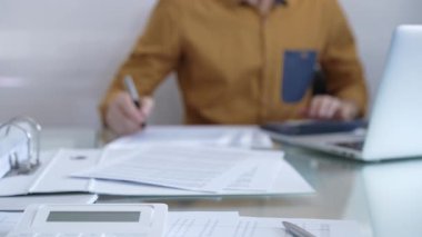 Hesap makinesi, büyüteç, kalem ve belgeler muhasebecilerin masasında. Sarı tişörtlü erkek muhasebeci, arka planda belgelerle çalışıyor. İş ve denetim kavramı.