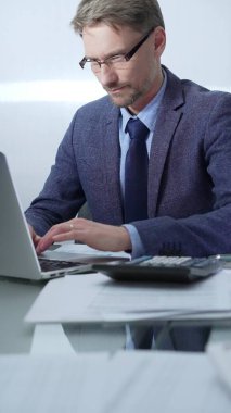 Mavi takım elbiseli iş adamı ofis setindeki cam masada dizüstü bilgisayar, hesap makinesi ve belgelerle çalışıyor. Düşük ışıklandırma, dikey portre. İş adamları ve denetim.