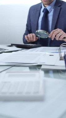 Mavi takım elbiseli bir işadamı büyüteçle mali raporları analiz ediyor ve profesyonel ofis ortamında hesap makinesi kullanıyor. Düşük ışıklandırma, yakın dikey görüntü. İş adamları, denetim.