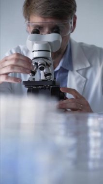 Laboratuvar önlüğü giyen ve koruyucu gözlük takan erkek bilim adamı mikroskop kullanarak modern laboratuvar düzeninde araştırma yapıyor, portre görüntüsü çiziyor. Çaktırmadan ışıklandırma videosu. Bilim ve Tıp.