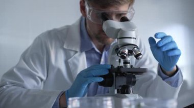 Mavi eldiven giyen, koruyucu gözlüklü erkek araştırmacı modern laboratuarda mikroskop kullanıyor, portre görüntüsü. Çaktırmadan ışıklandırma videosu. Bilim ve Tıp kavramları.