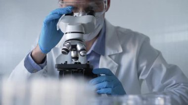 Mavi eldiven, koruyucu gözlük ve tıbbi maske takan bilim adamı araştırmacı modern laboratuarda mikroskop kullanıyor, portre görüntüsü. Çaktırmadan ışıklandırma videosu. Bilim ve Tıp.