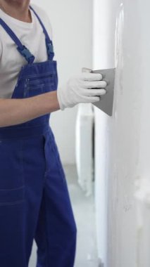 Meçhul inşaat işçisi beyaz eldiven ve mavi tulum giyen kadın, macun bıçağı kullanarak beyaz bir duvara alçı sürüyor. Ev yenileme projesi sırasında pürüzsüz bir yüzey oluşturuyor.