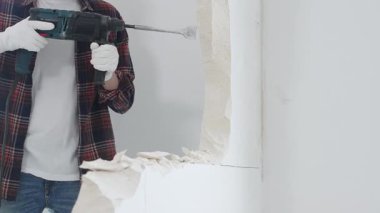 Kırmızı kareli kot pantolon ve koruyucu eldiven giyen kimliği belirsiz erkek inşaat işçisi, döner çekiç matkabıyla beyaz duvarı yıkıyor, toz üretiyor, yakın görüş sağlıyor. Yenileme konsepti.