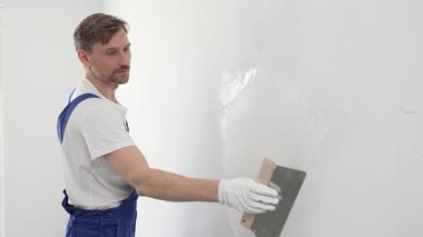 Koruyucu eldivenler ve mavi inşaat tulumları giyen erkek inşaat işçisi, tadilat projesi sırasında profesyonel alçı levha bantlı bıçak kullanarak duvarlara sorunsuzca alçı yayıyor. Portre