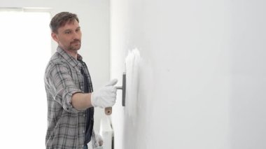 Eldiven ve ekose gömlek giyen orta yaşlı inşaat işçisi, yenileme projesi sırasında pürüzsüz bir yüzey oluşturmak için beyaz duvar alçılamak için son malayı kullanıyor..